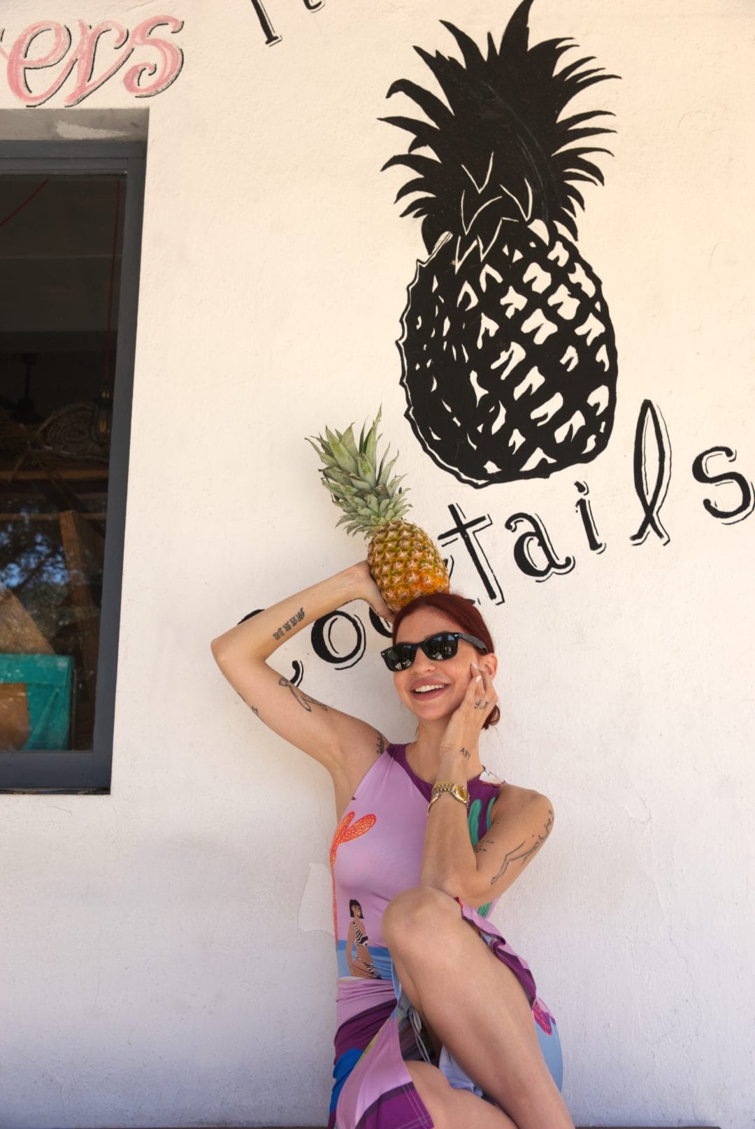 Woman holding a pineapple against a wall with a pineapple design and 'tails' text.
