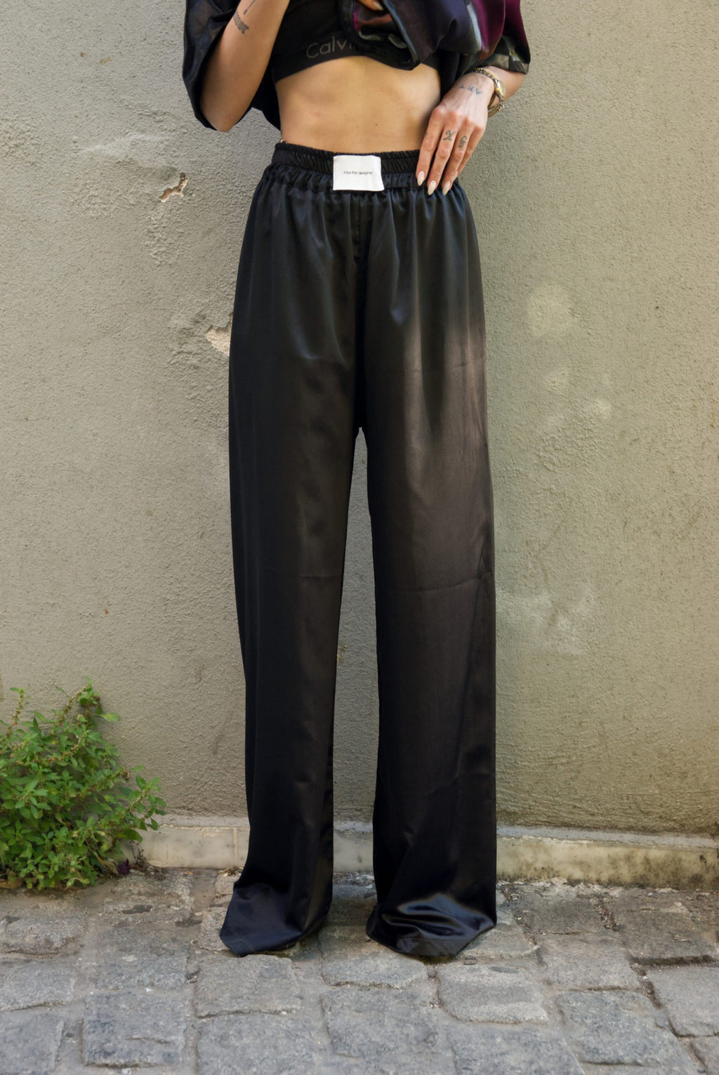 Person wearing black satin pants standing against a beige wall.