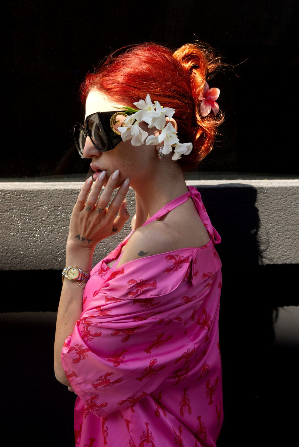 Woman with red hair wearing sunglasses and a pink floral dress against a black background