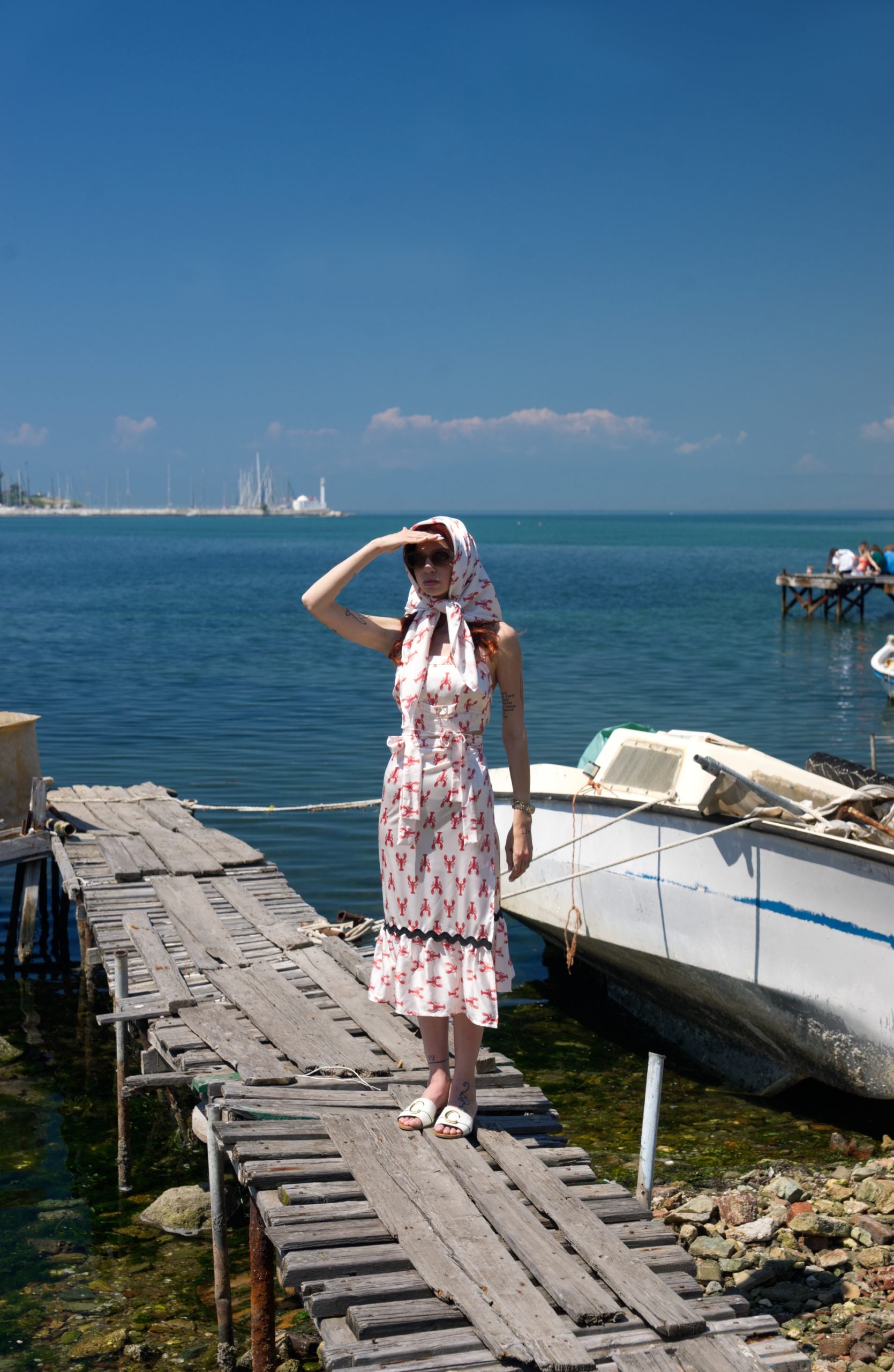 Woman in a dress standing on a wooden dock by the water with a clear blue sky.