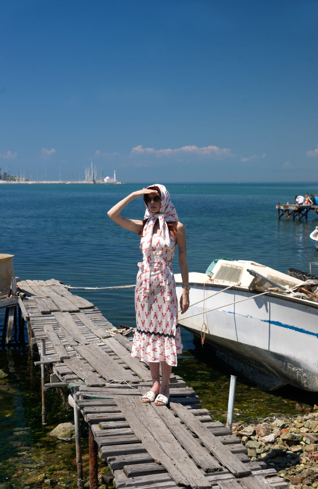 Woman in a dress standing on a wooden dock by the water with a clear blue sky.