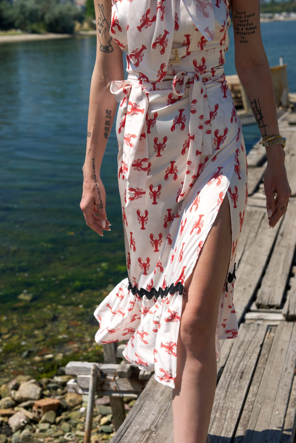Person wearing a white dress with red lobster pattern on a wooden dock by a body of water.
