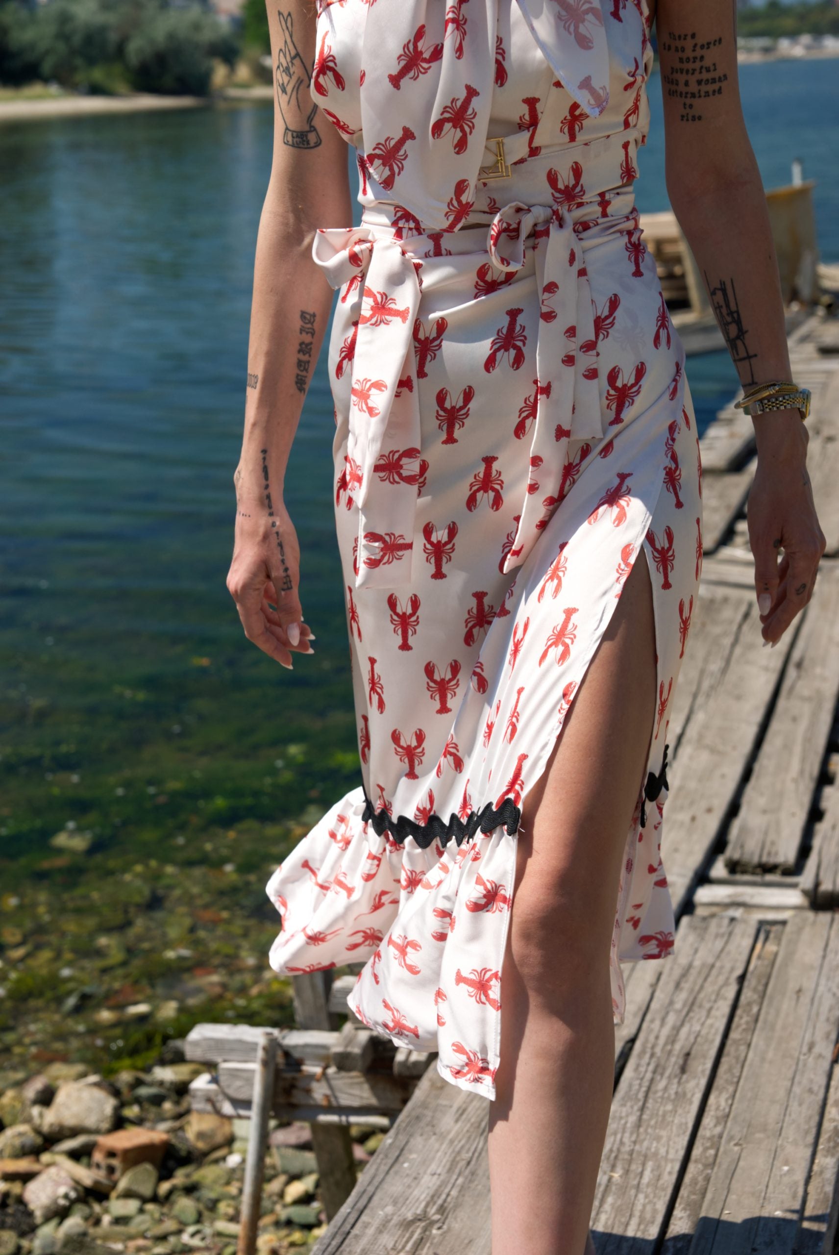 Person wearing a white dress with red lobster pattern on a wooden dock by a body of water.