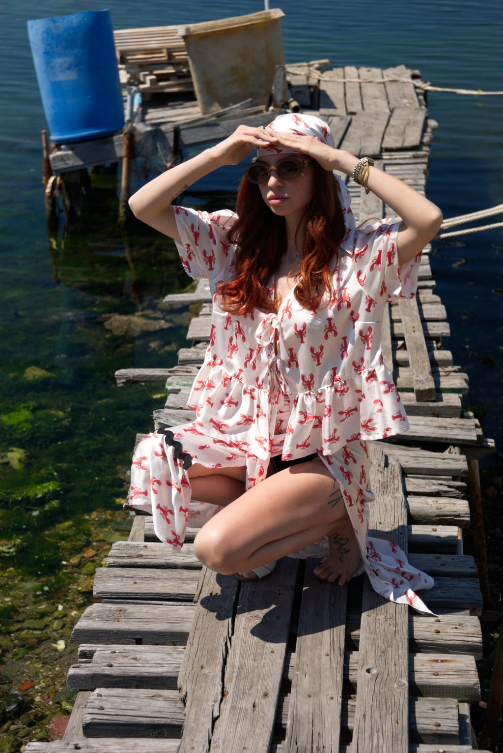 Woman in a white dress with red patterns sitting on a wooden dock by a body of water.