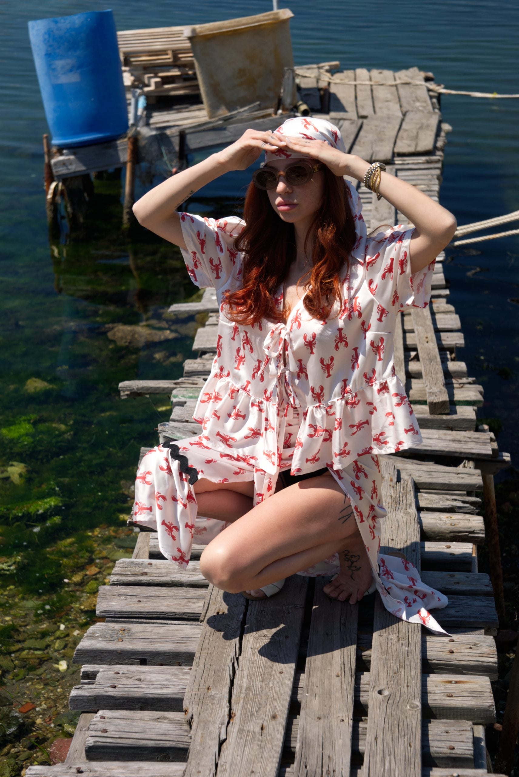 Woman in a white dress with red patterns sitting on a wooden dock by a body of water.