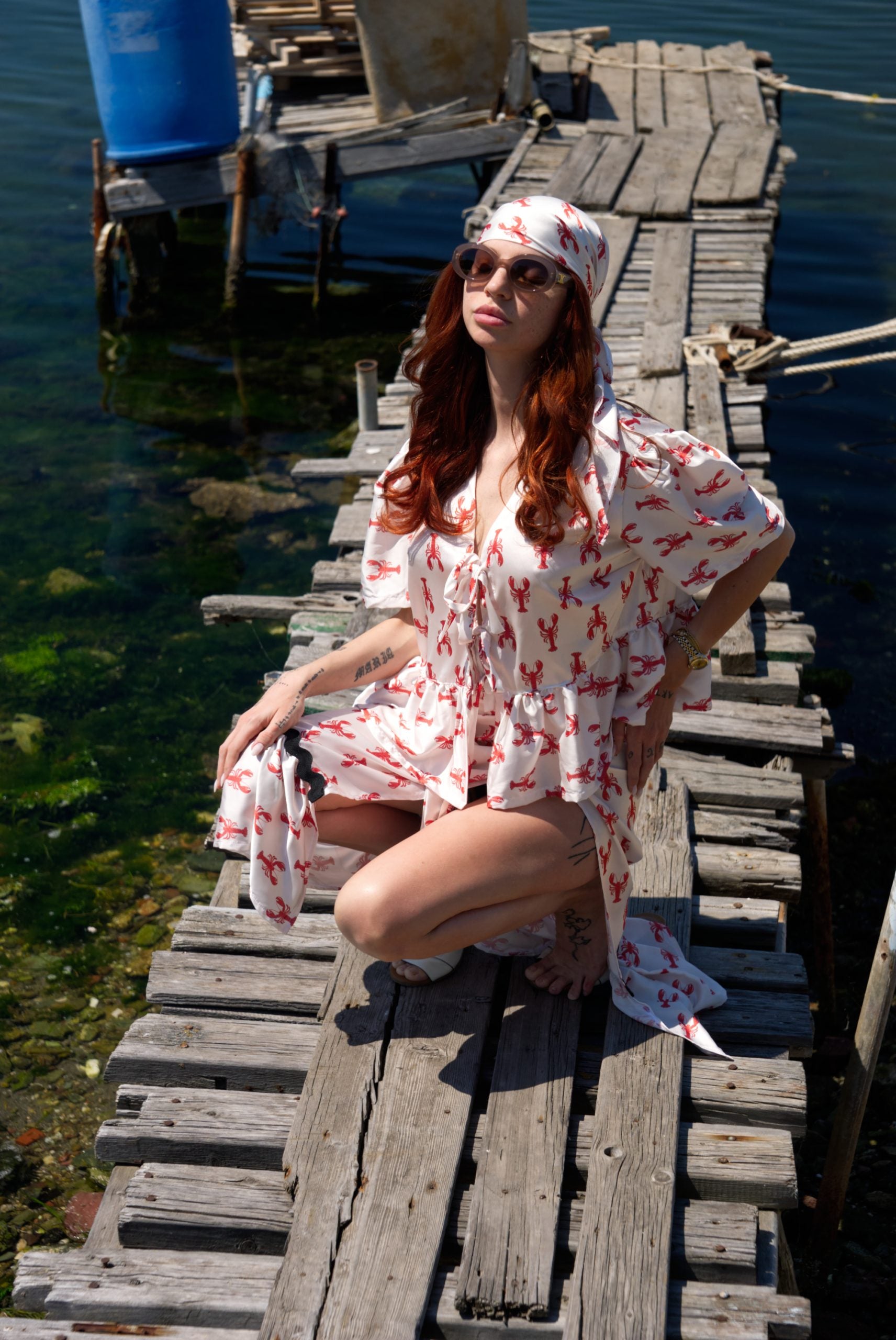 Woman in a red and white patterned dress sitting on a wooden dock by the water.