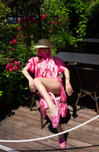 Woman in a pink dress and hat sitting outdoors with flowers and furniture in the background