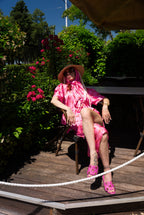 Person in a pink dress and hat sitting on a wooden deck with greenery in the background