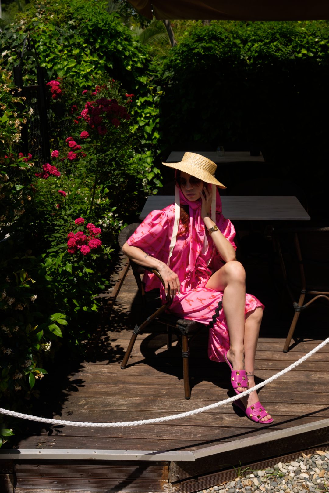 Woman in a pink dress and straw hat sitting on a wooden deck with greenery around.