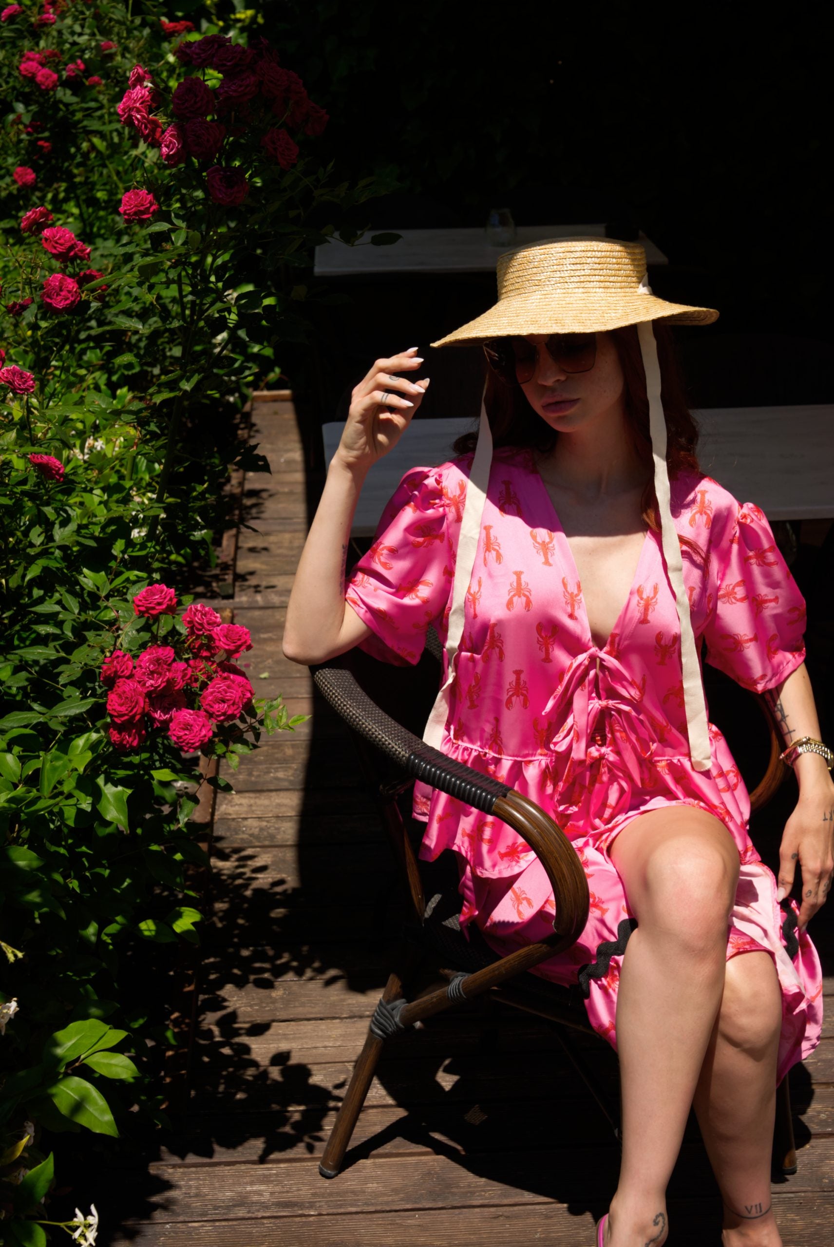 Woman in a pink dress and straw hat sitting on a wooden chair in a garden.