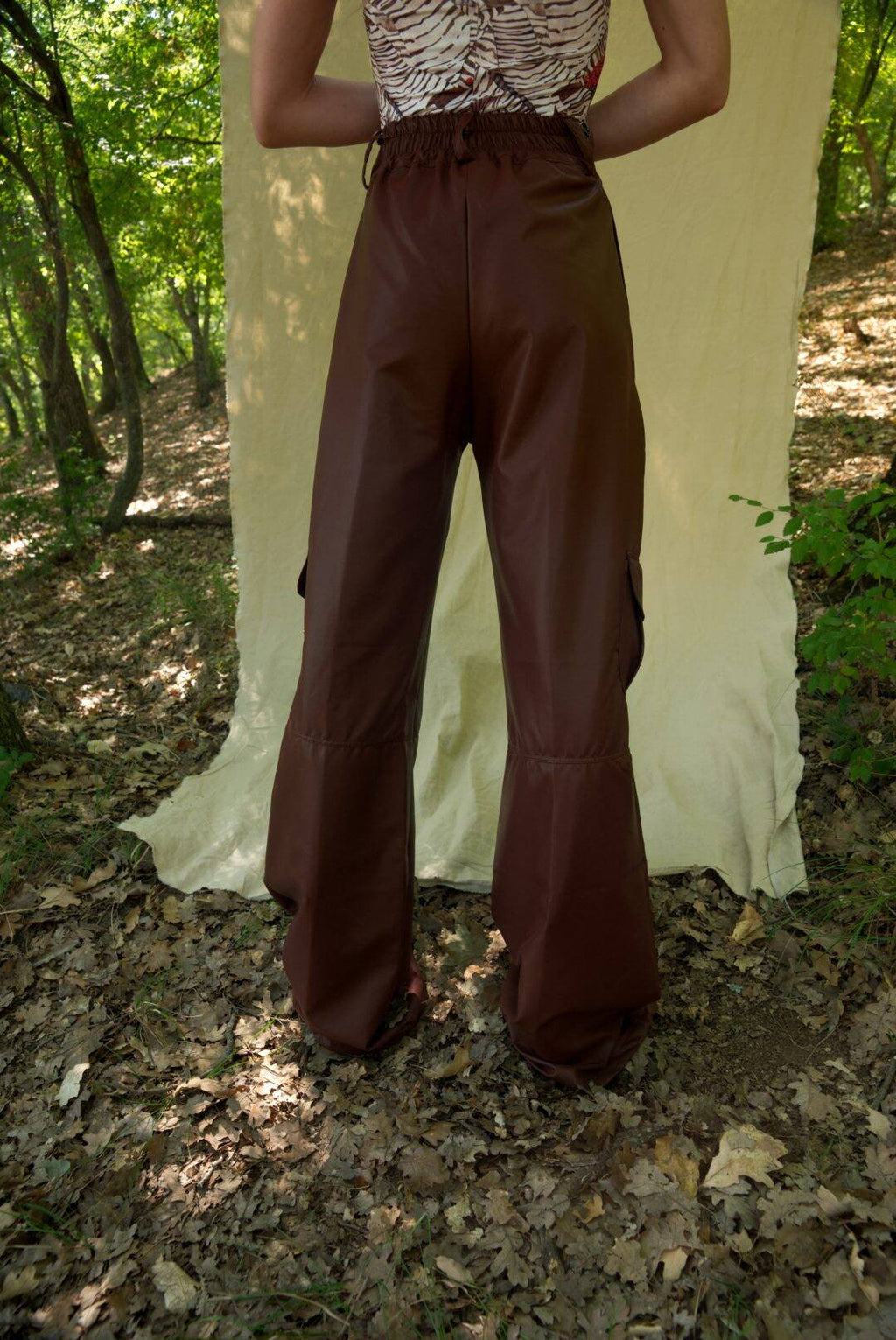 Person wearing brown bell-bottom pants standing in a forest setting with a white curtain.