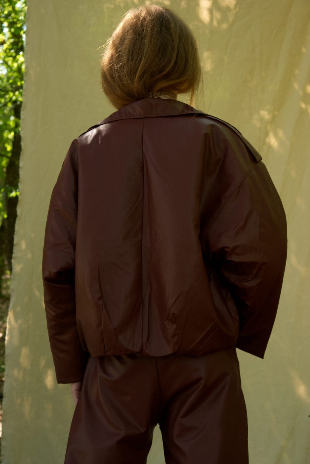Person wearing a brown jacket and pants standing against a white wall with greenery.