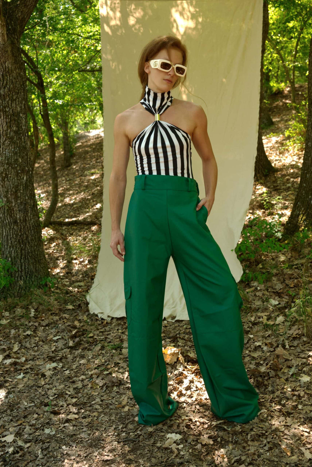 Woman in a black and white striped top and green pants standing in a forest setting.