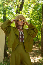 Person in a green outfit with a fur-lined hood and sunglasses in a forest setting