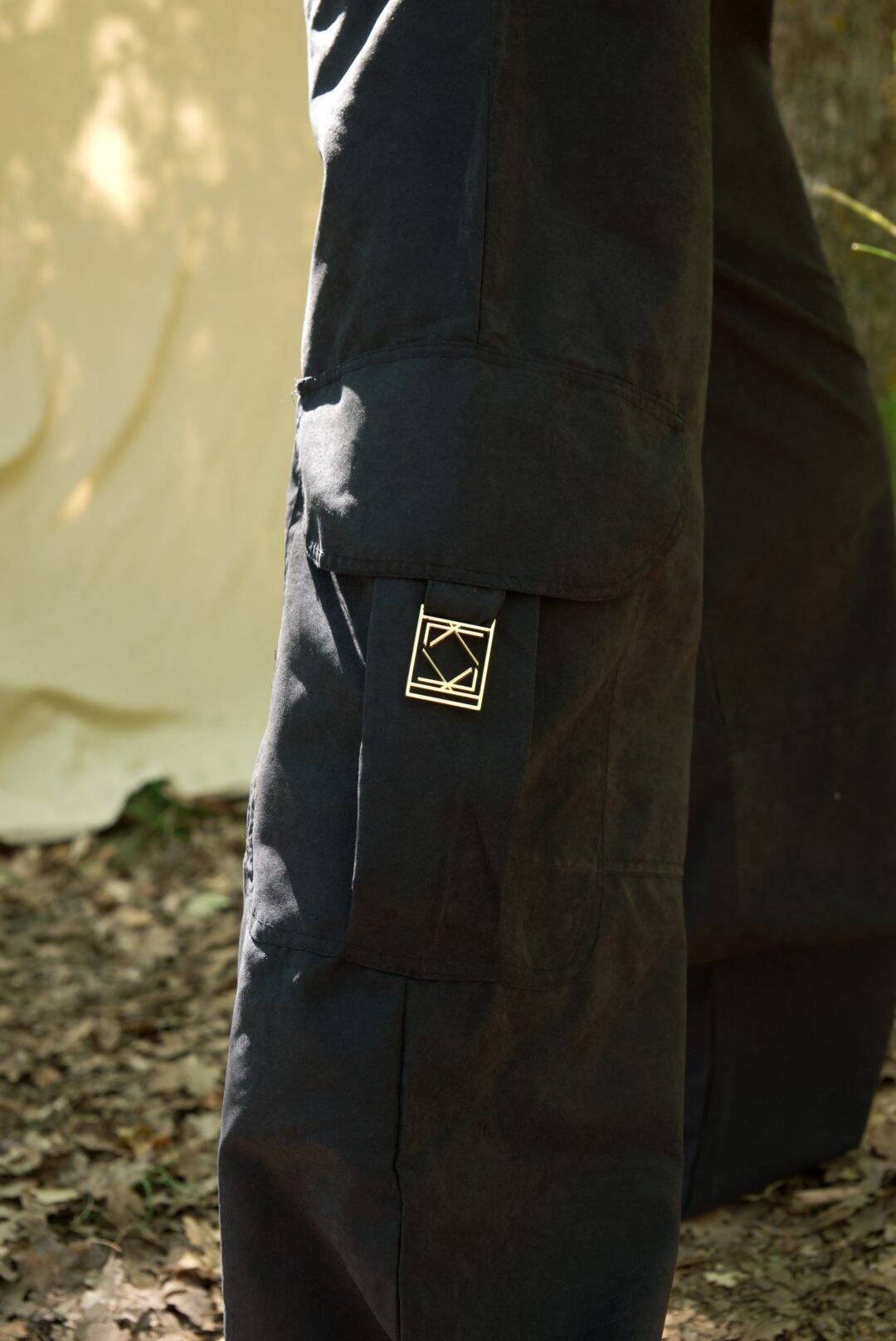 Close-up of dark pants with a brand logo on the pocket against a natural background.
