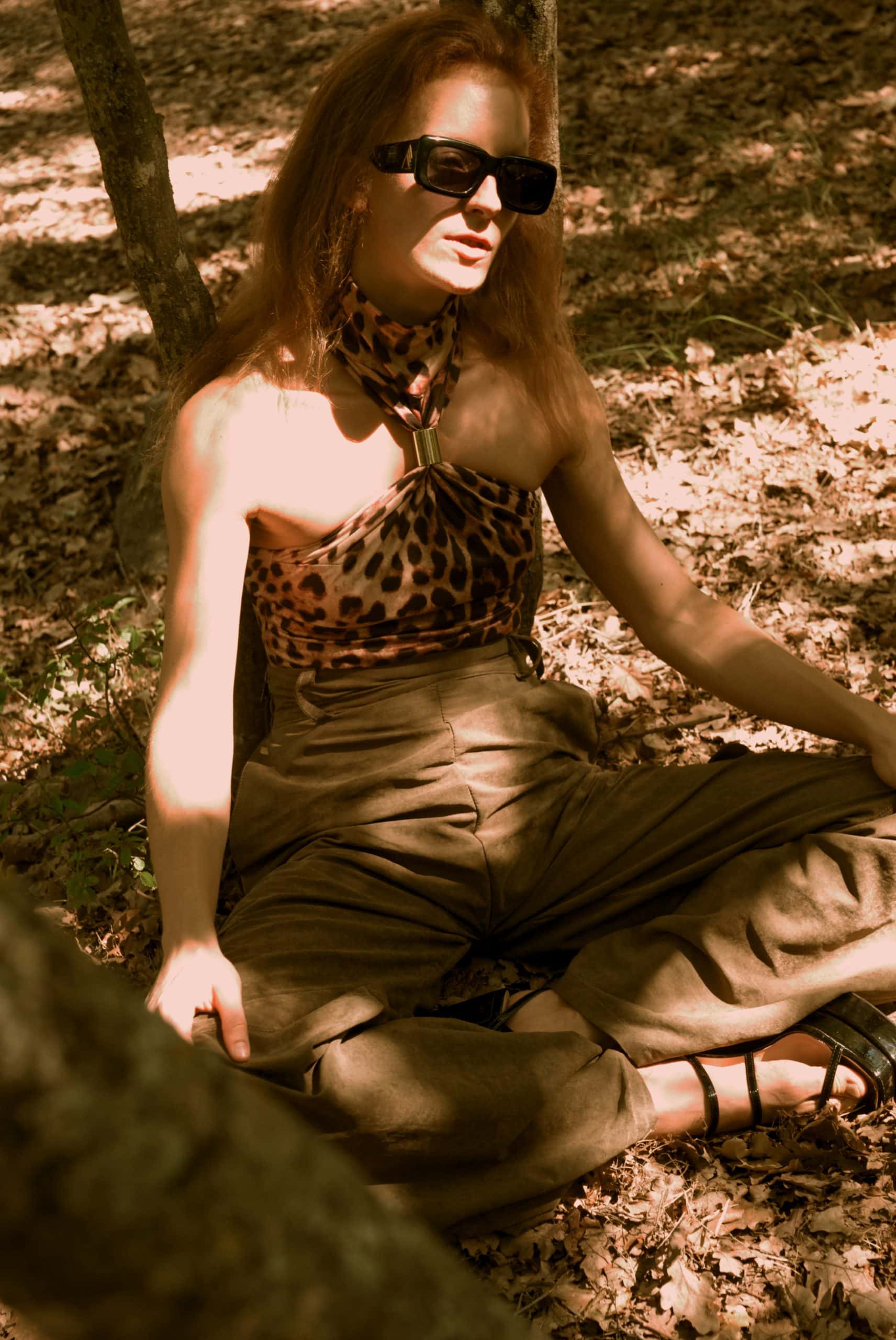Woman sitting on the ground wearing a leopard print top and brown pants in a natural setting.