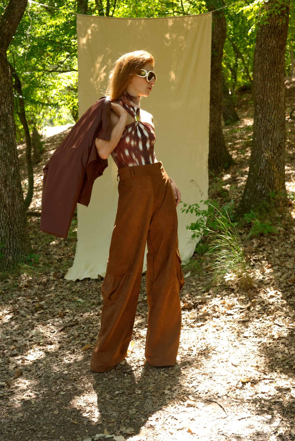 Woman in a forest setting wearing a patterned top and brown wide-leg pants.