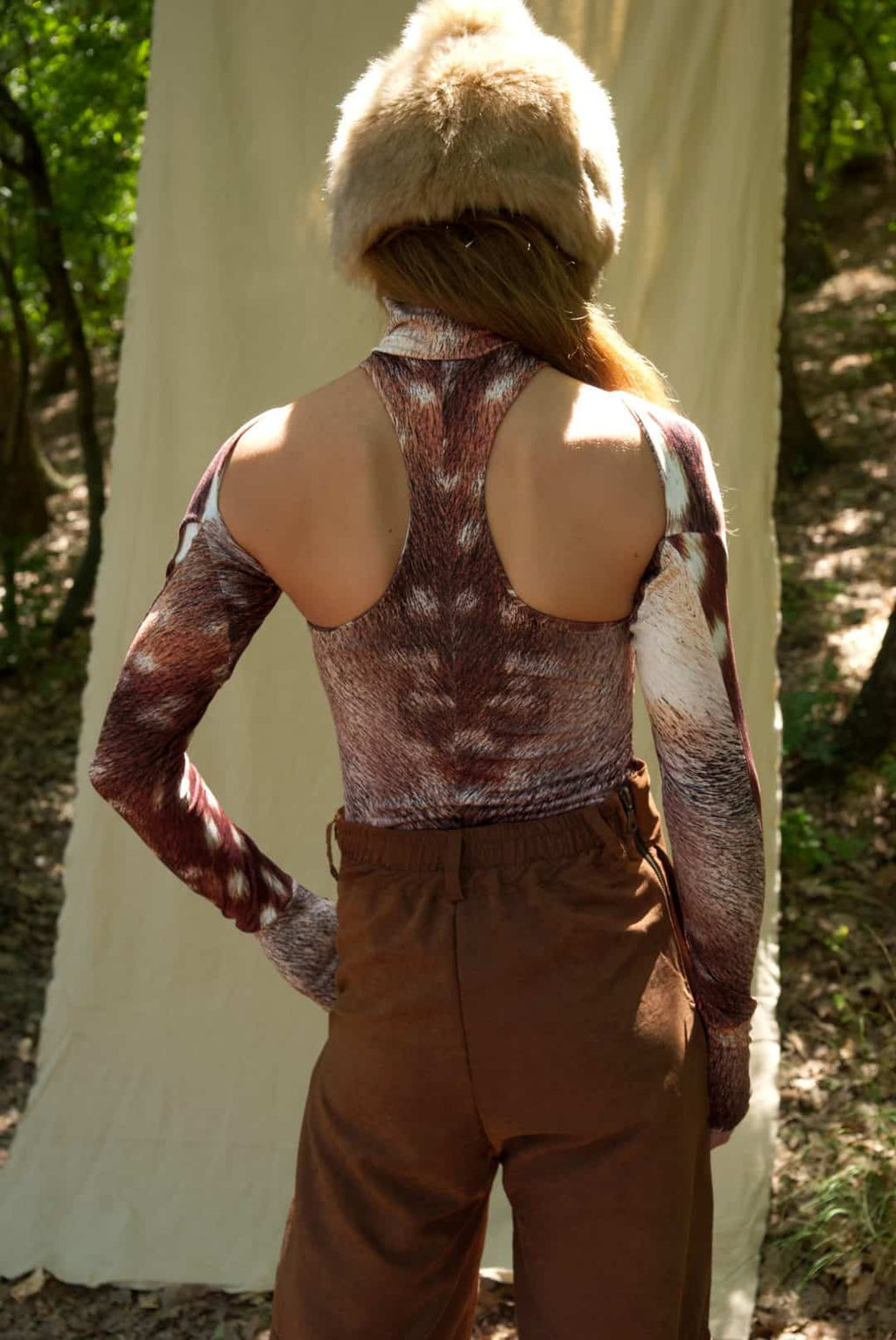 Person wearing a patterned top and brown pants with a fur hat in a natural setting.