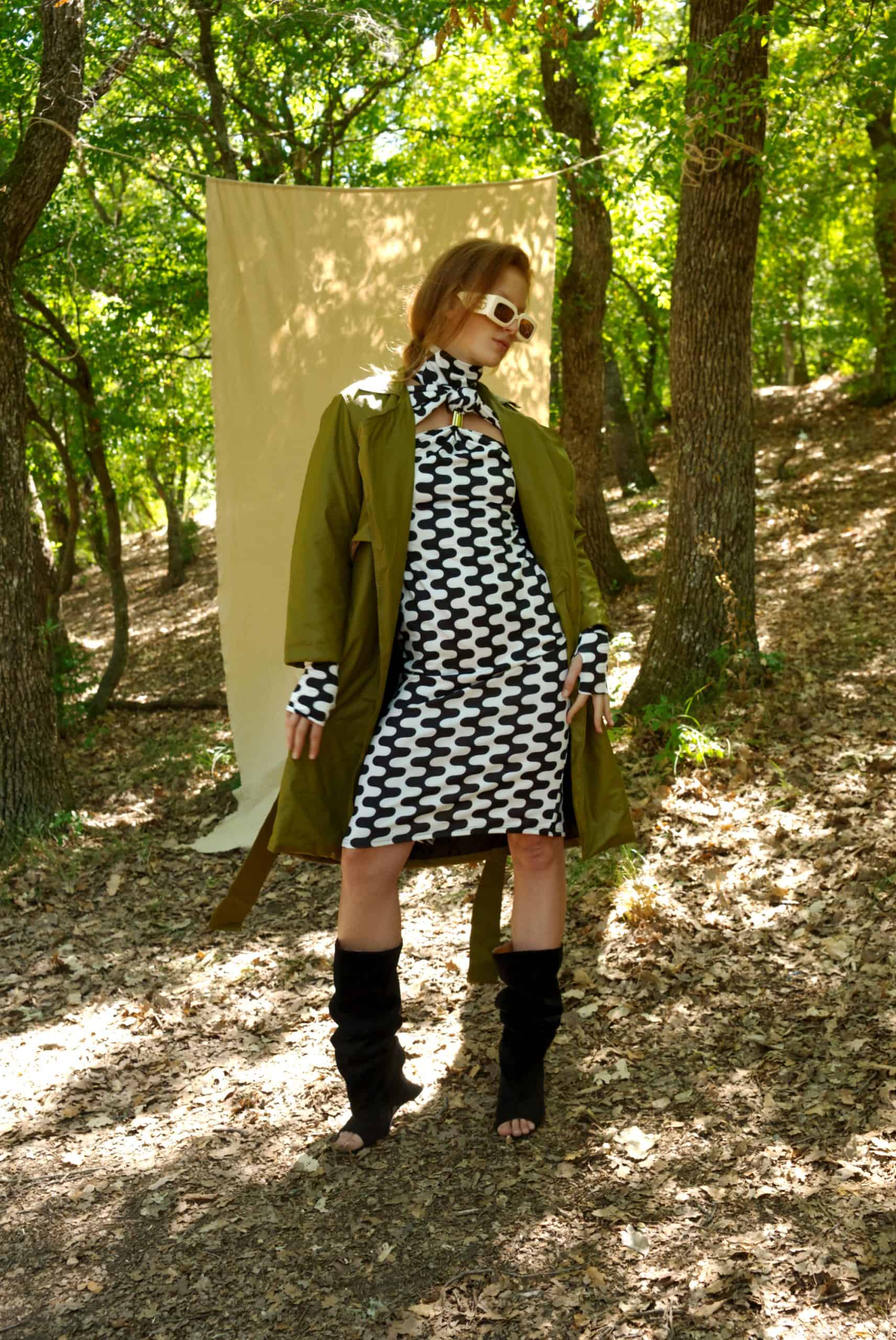 Woman in a green coat and black and white dress standing in a forest.
