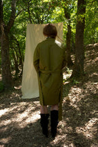 Person wearing a green trench coat standing in a forest with a white sheet draped over a tree branch.