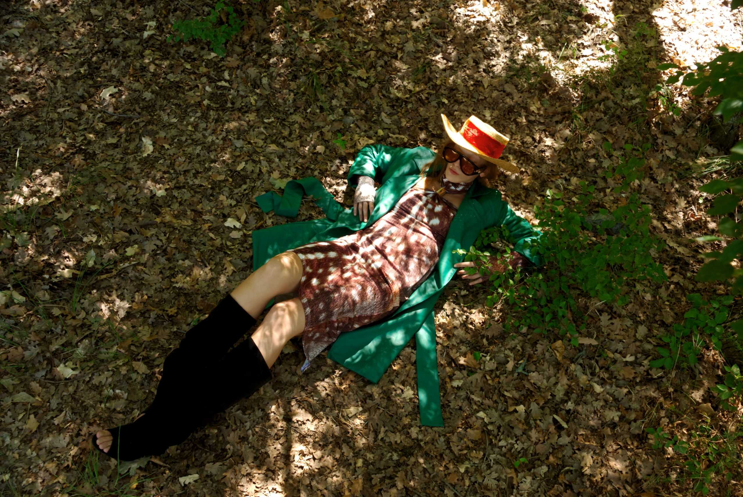 Person lying on the ground wearing a green dress, patterned top, and black shoes with a hat.