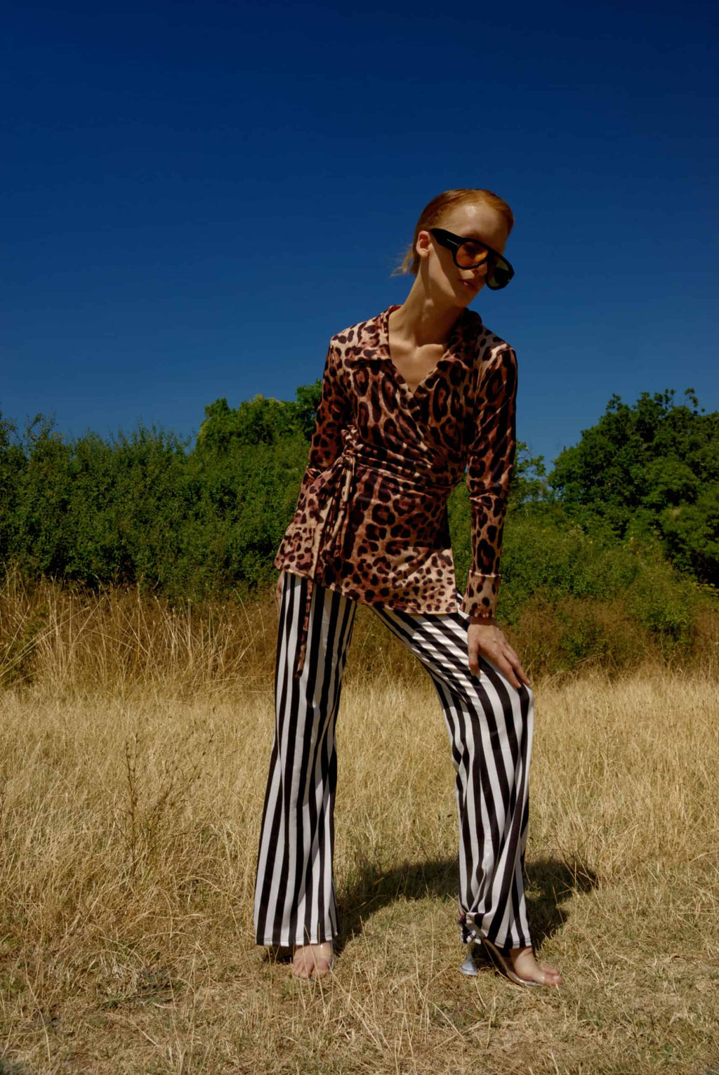 Person wearing a leopard print top and black and white striped pants standing in a field with a blue sky.