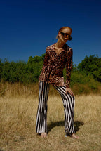 Person wearing a leopard print top and black and white striped pants standing in a field with a blue sky.