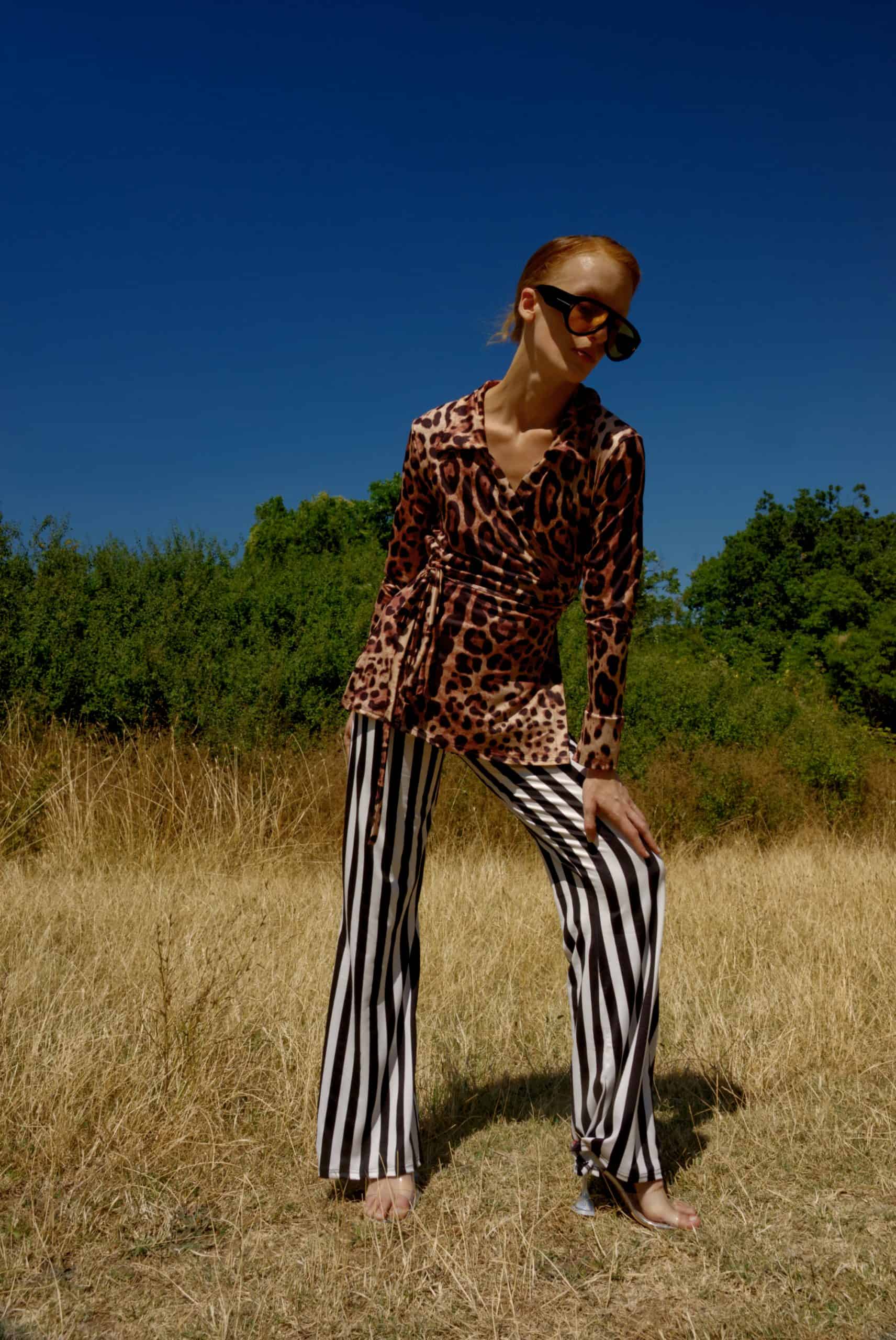 Person wearing a leopard print top and black and white striped pants standing in a field with a blue sky.