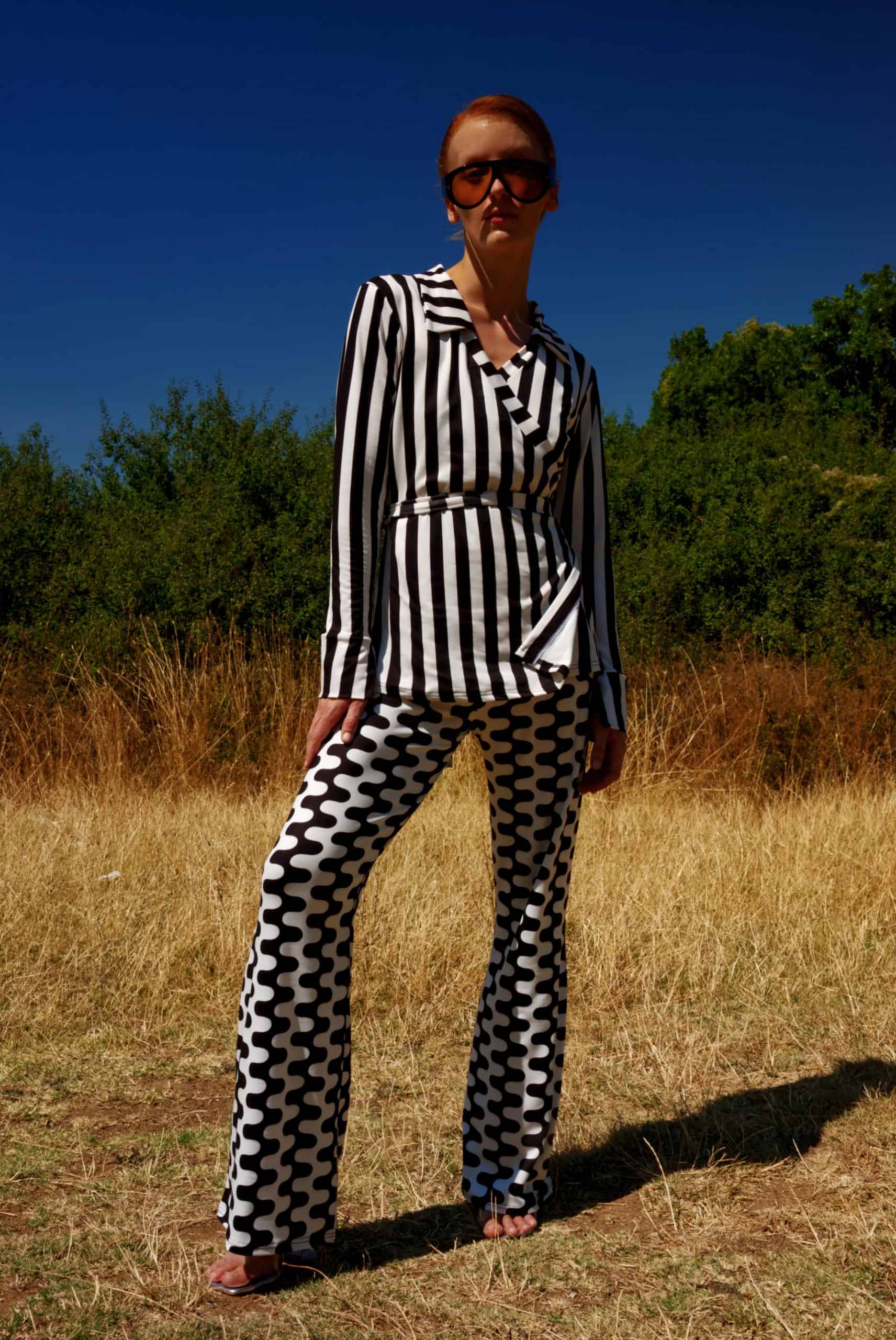 Woman in a black and white striped top and patterned pants standing in a field with trees in the background.