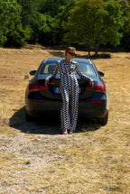 Person in a checkered outfit standing next to a car in a field with trees in the background