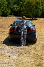 Person in a checkered outfit standing next to a car in a field with trees in the background