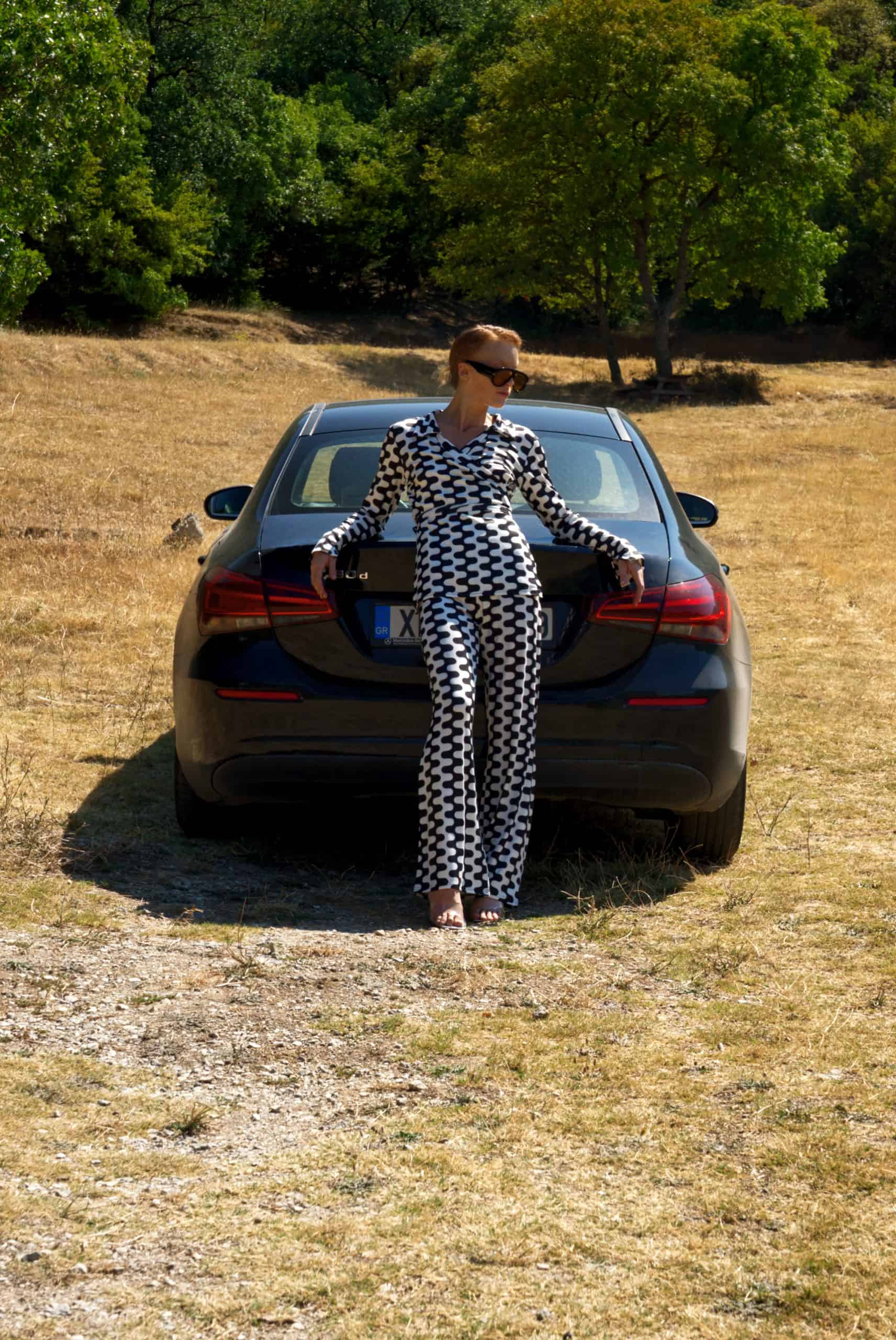 Person in a checkered outfit standing next to a car in a field with trees in the background