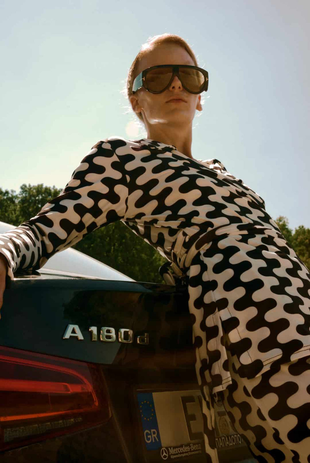 Person wearing sunglasses and a patterned outfit leaning against a car with 'A180' branding.
