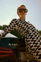 Person wearing sunglasses and a patterned outfit leaning against a car with 'A180' branding.