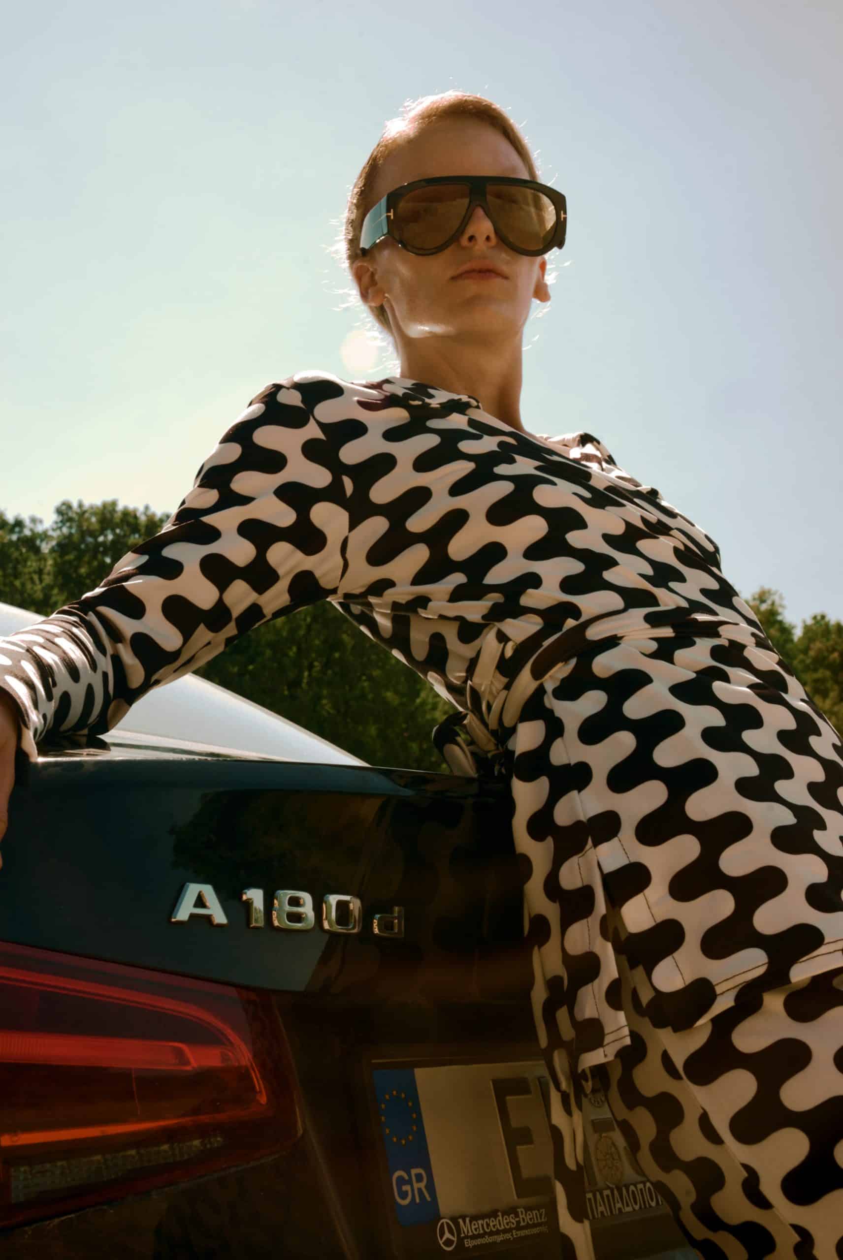 Person wearing sunglasses and a patterned outfit leaning against a car with 'A180' branding.