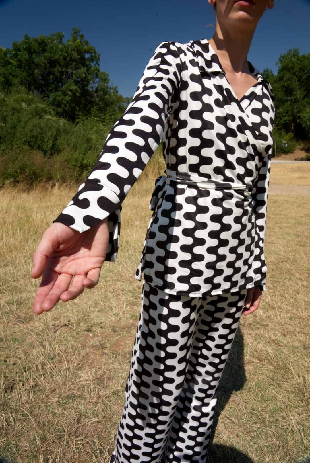 Person wearing a black and white patterned outfit standing in an outdoor setting with trees and grass.