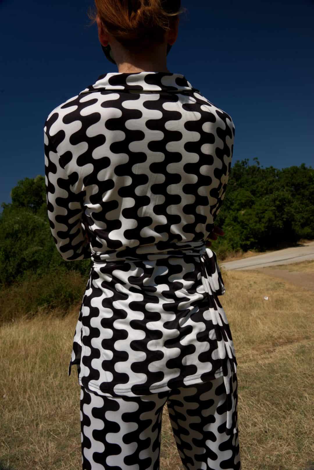 Person wearing a black and white patterned outfit standing in an open field with trees in the background.