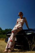 Woman in a patterned outfit sitting on a car in a field with a clear blue sky.