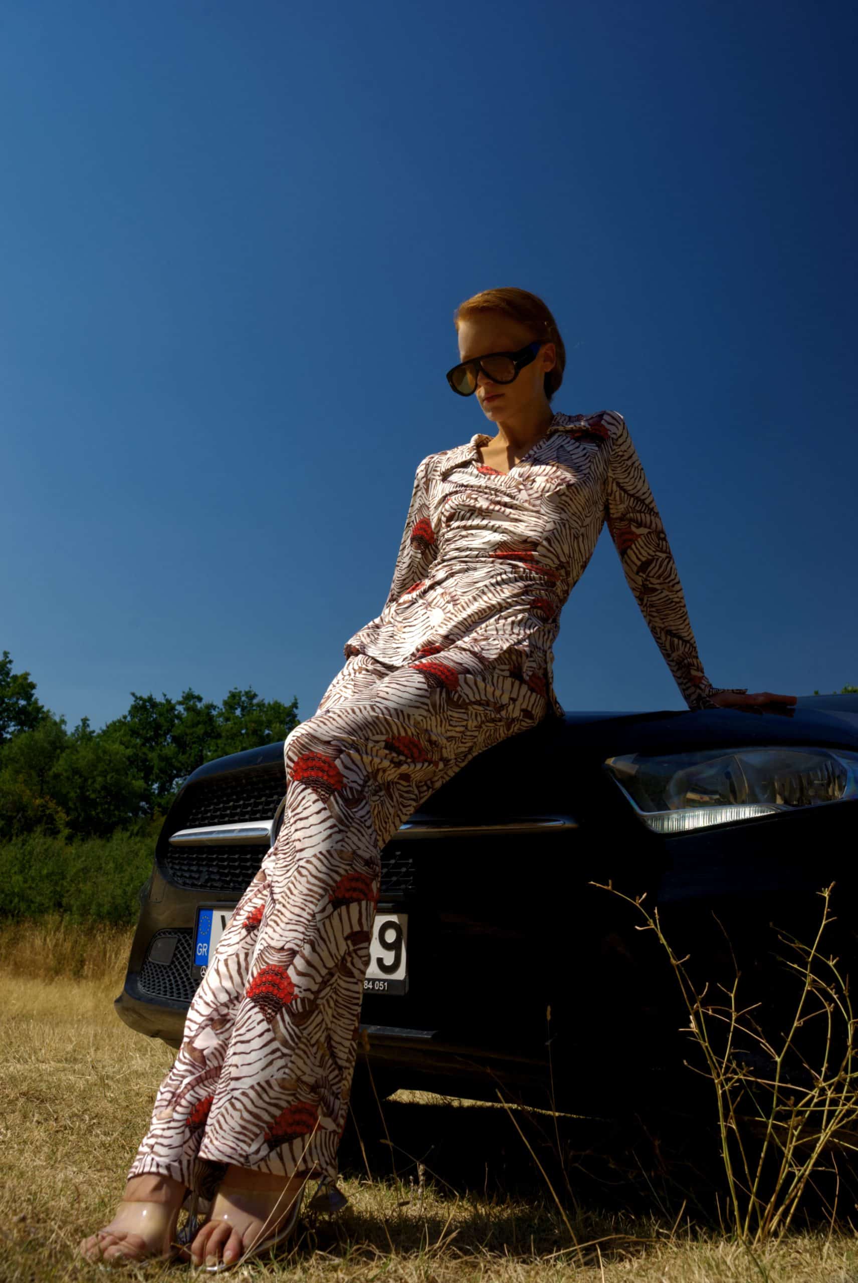 Woman in a patterned outfit sitting on a car in a field with a clear blue sky.