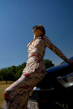 Person in a patterned outfit leaning against a car with a clear blue sky and trees in the background