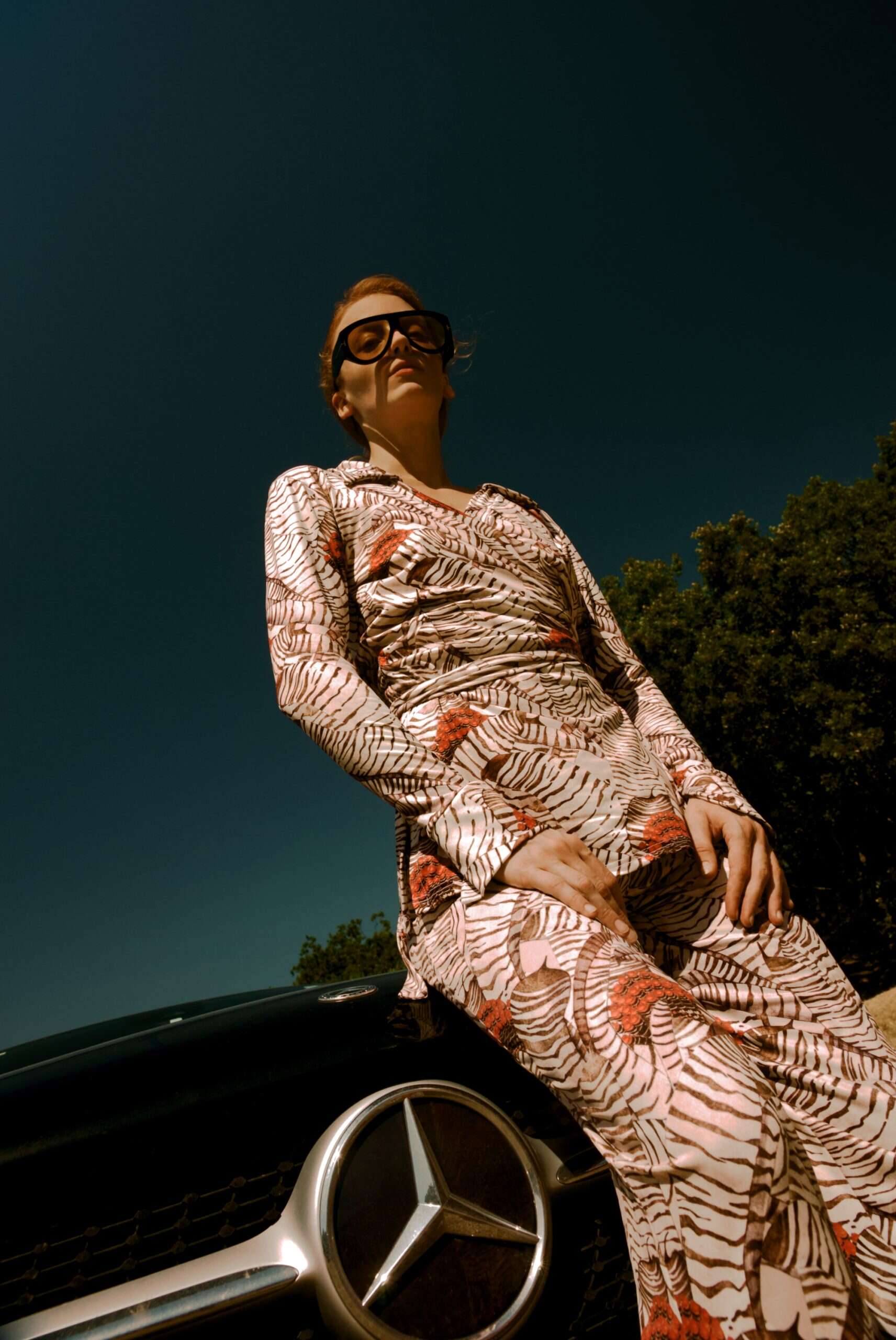 Person in a patterned outfit leaning against a car with a clear sky background