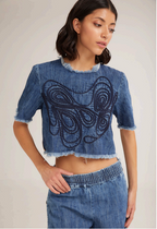 A female model wearing high-waisted denim pants with a large front pocket and an embroidered design on the chest area. She is also wearing a denim top with a large front pocket.