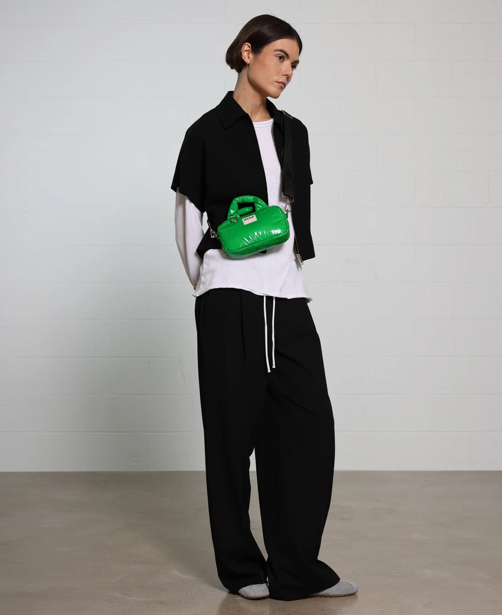 Person wearing a black jacket, white shirt, and black pants with a green bag on a plain background