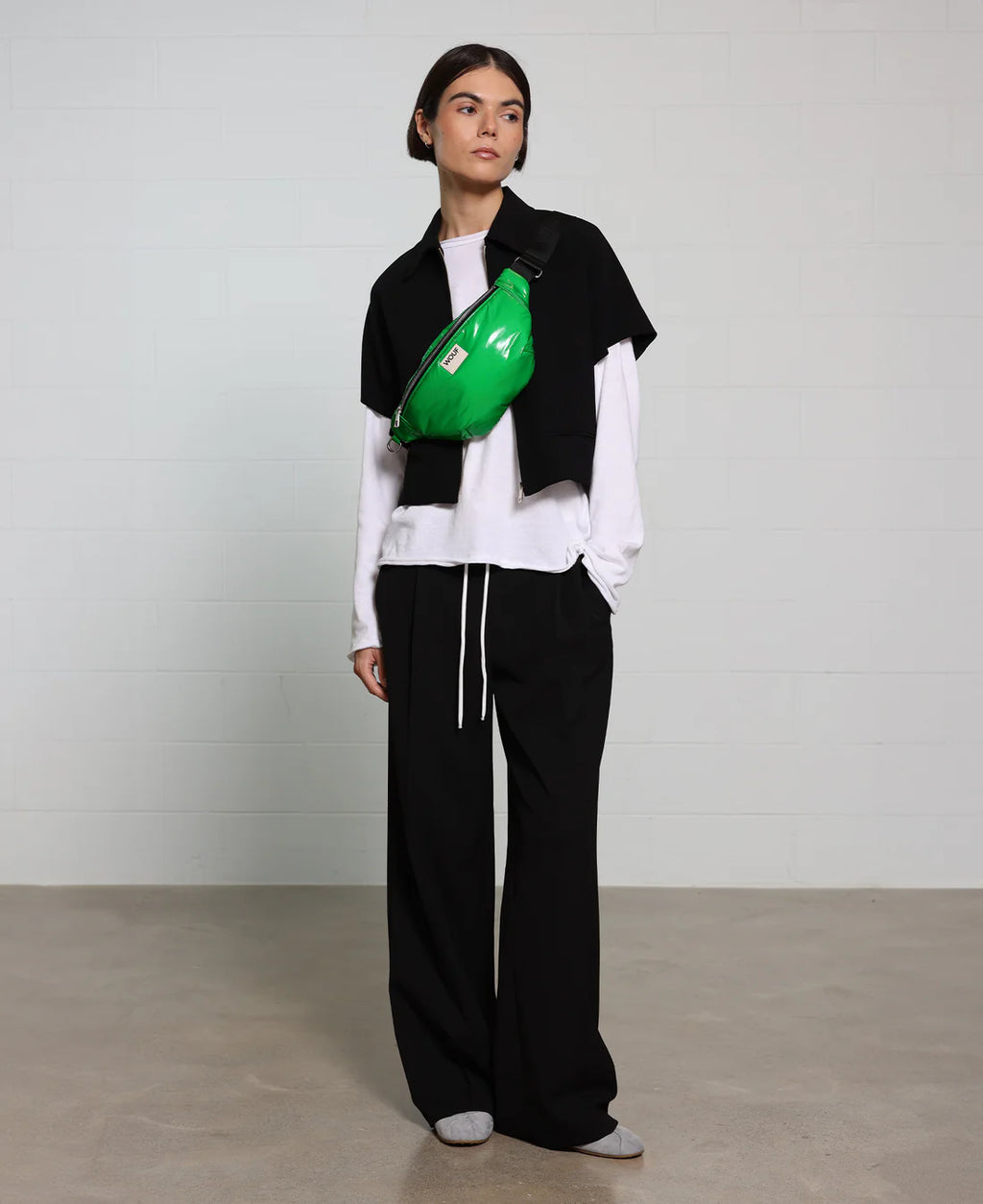 Person wearing a black vest, white shirt, and black pants with a green bag over one shoulder against a plain background.