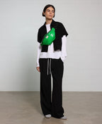 Person wearing a black vest, white shirt, and black pants with a green bag over one shoulder against a plain background.