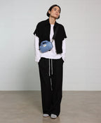 Person wearing a black vest, white shirt, and black pants with a blue bag, standing against a plain background.
