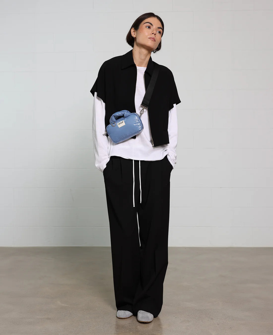 Person wearing a black vest, white shirt, and black pants with a blue bag, standing against a plain background.