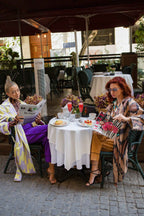 Two women at an outdoor cafe table, reading magazines and newspapers, with desserts and drinks served on a white tablecloth.