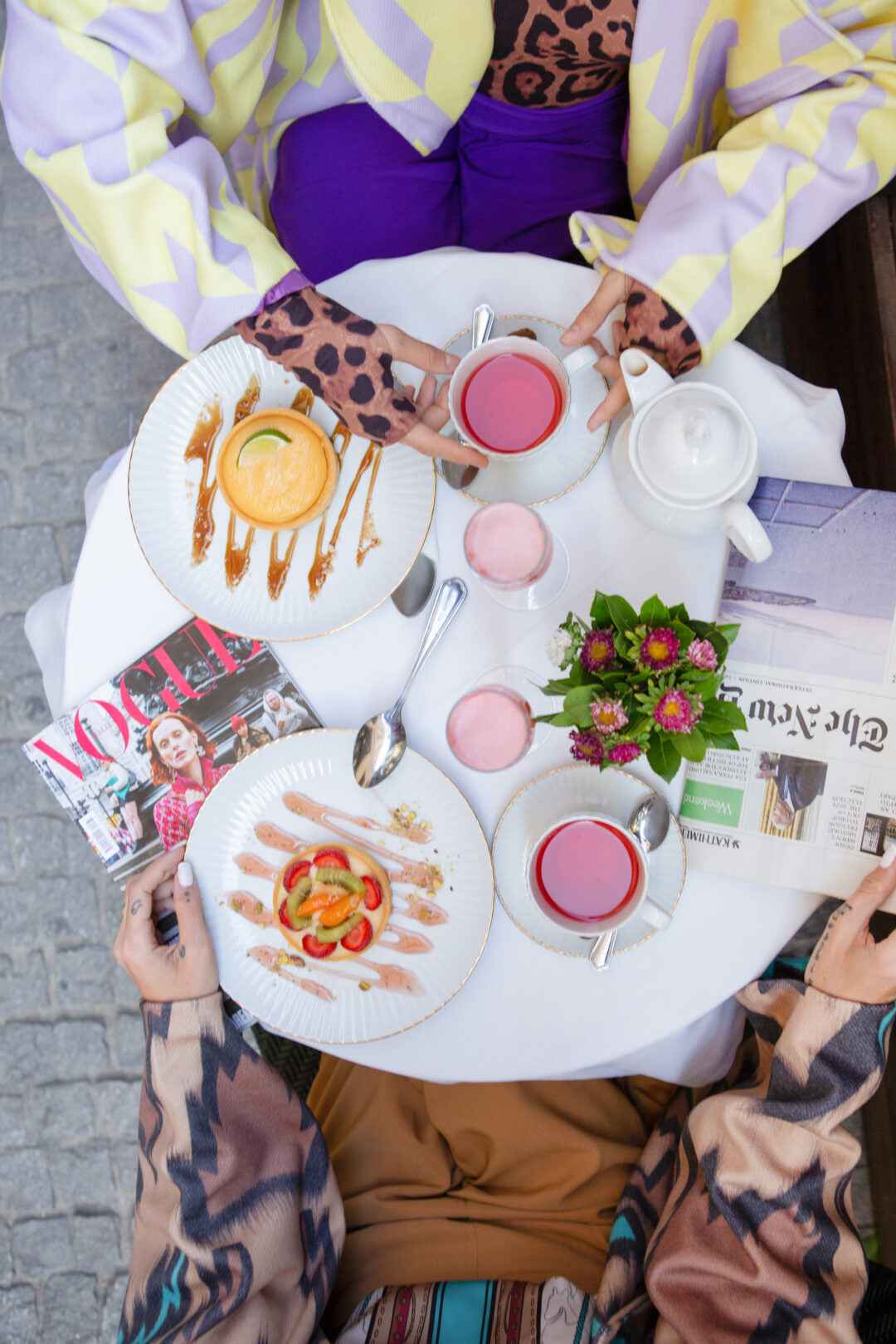 Two people at a table with pastries, tea, and magazines. One wears a yellow and purple jacket, the other a brown and turquoise jacket.