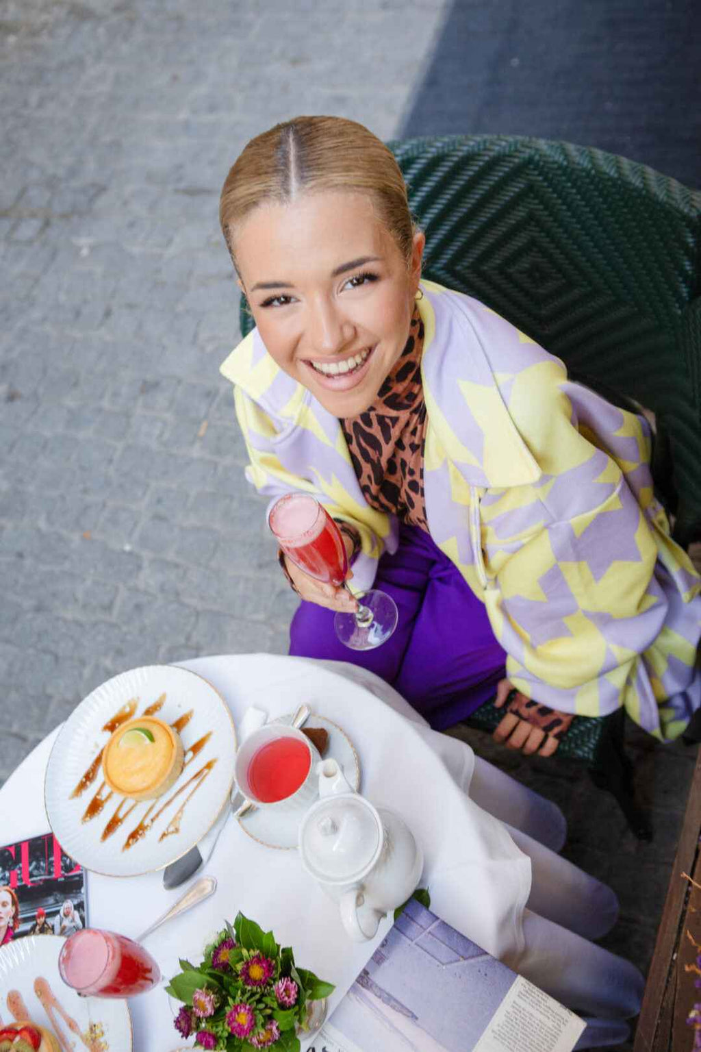 A woman in a patterned jacket holds a drink at a table with tea, a dessert, and flowers.